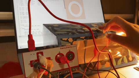 The image displays a hand interacting with the floral box interface under warm lamp light, pressing the wire loop buttons and triggering a glowing visual response on the laptop screen, using iron wire as the conductive input.