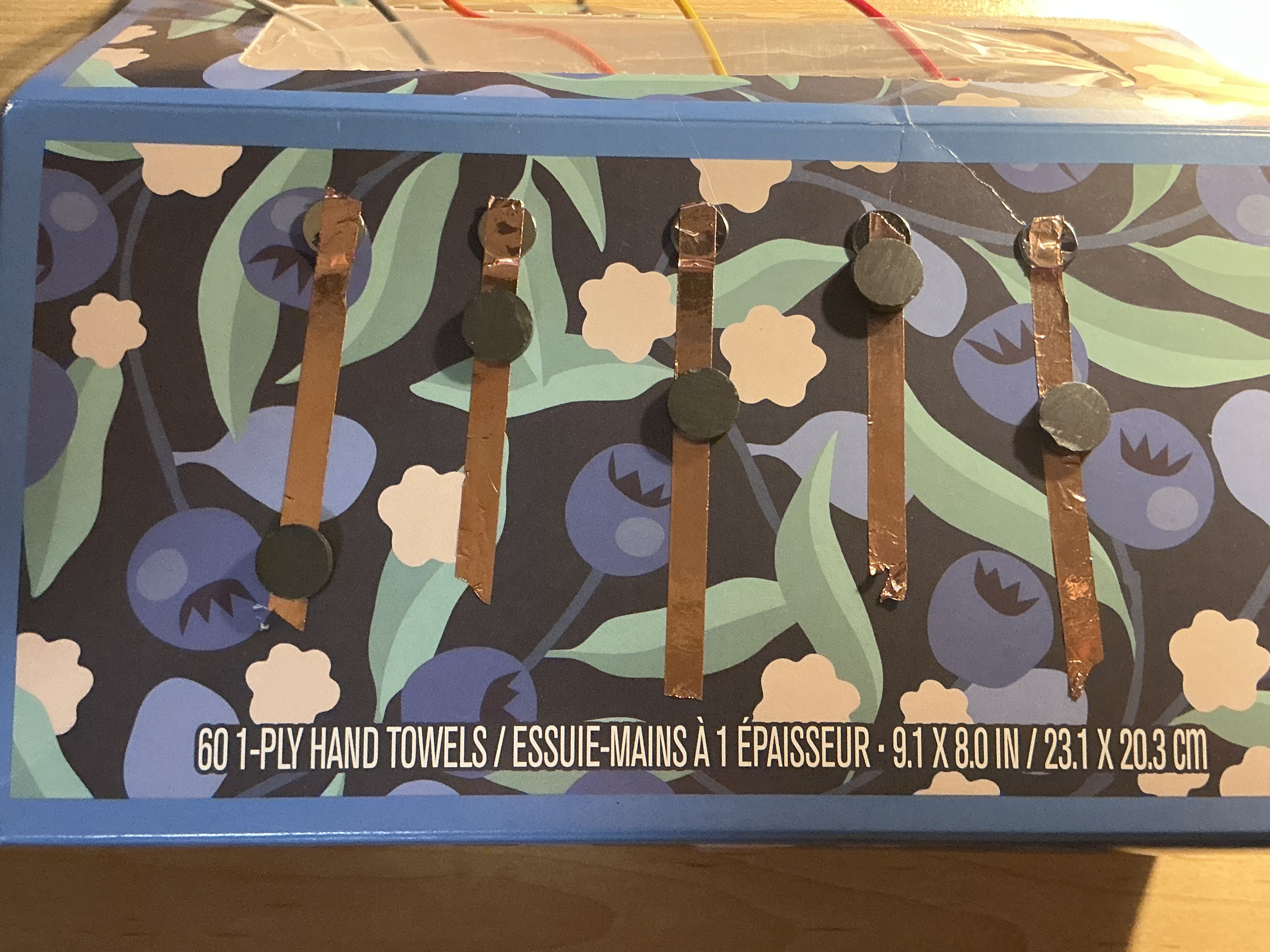 The image displays the blue floral box lying flat showing five copper tape strips each topped with a circular magnet aligned in a row, combining copper tape and magnets as a composite conductive button system.