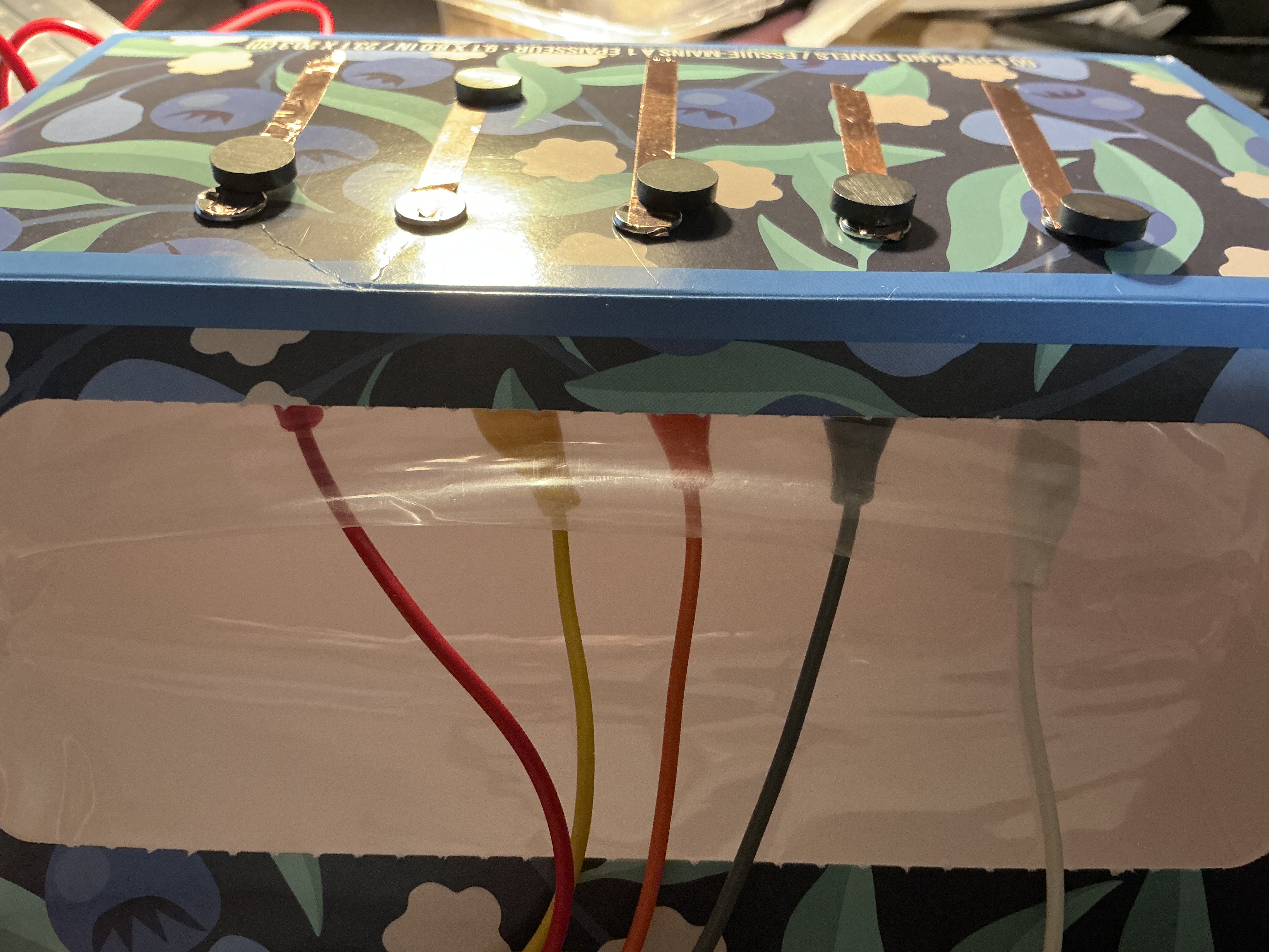 The image displays a close side-angle view of the blue floral box showing copper tape strips with circular magnets mounted on top and alligator clip wires exiting from below, using copper tape bridged with magnets as the conductive contact points.