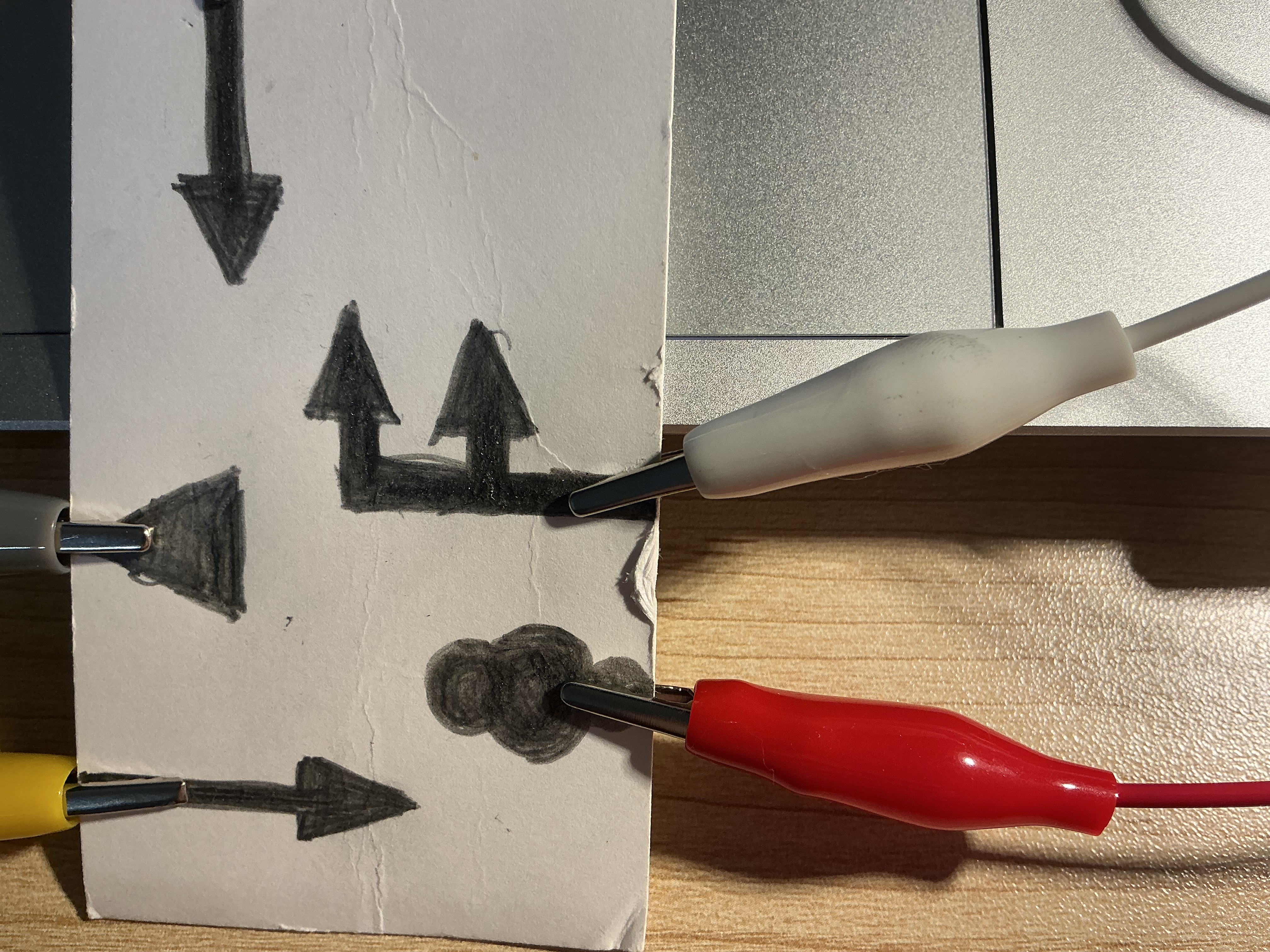 The image displays a close-up of the pencil-drawn card showing five directional arrow shapes with alligator clips biting into the thickest graphite-filled areas, using pencil graphite as the conductive trigger points.